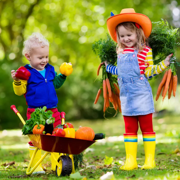 Two kids in colorful outfits are happily gardening, with one holding peppers and the other carrying carrots. A wheelbarrow is full of veggies.
