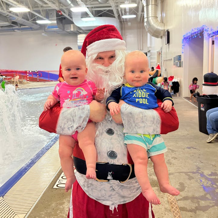 A person dressed as Santa is holding two infants in swimwear at a festive indoor pool setting.