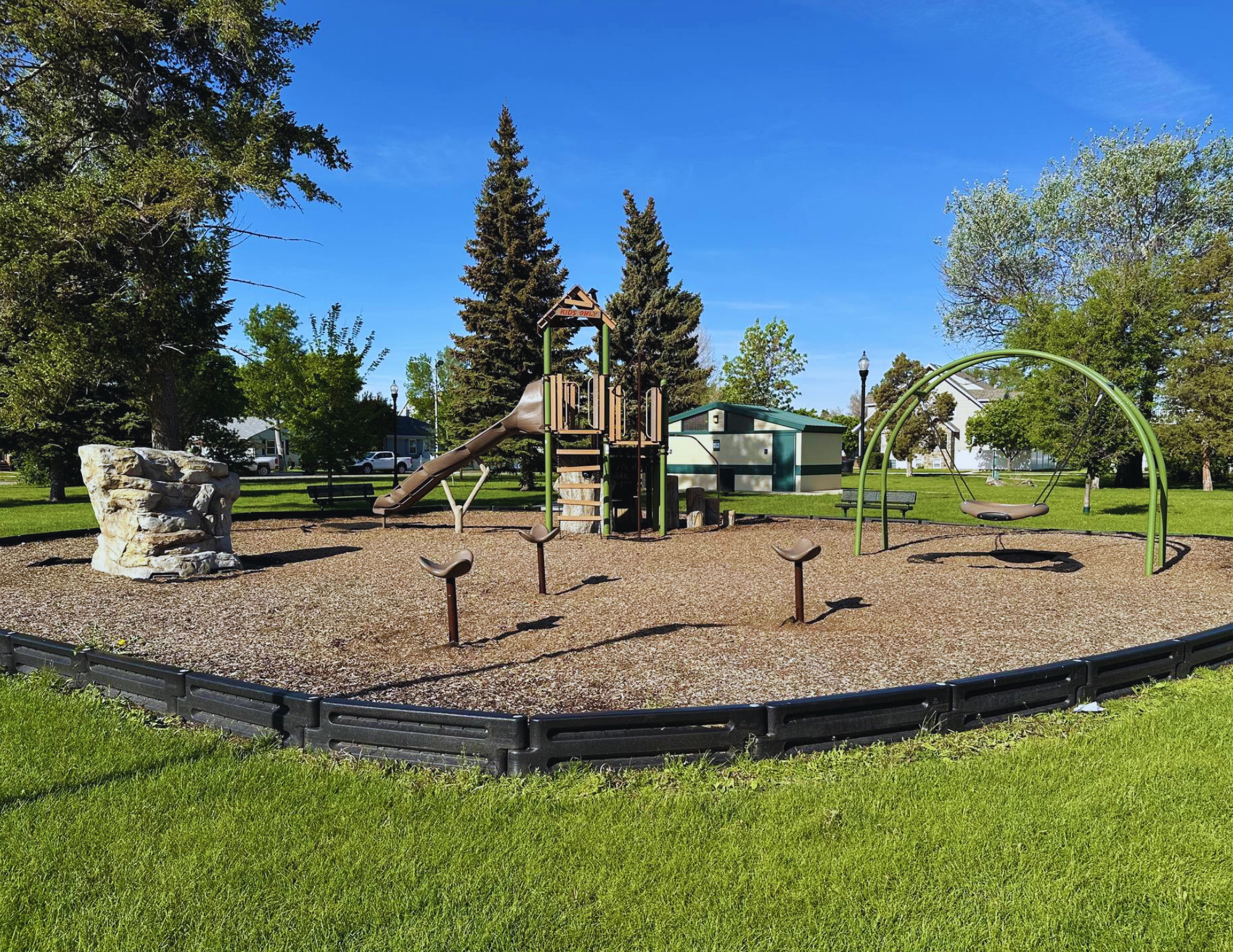 A playground with a slide, swings, climbing rock, and benches surrounded by trees and grass.