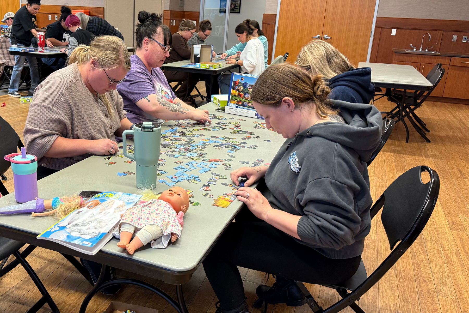 A group of women is assembling a puzzle at tables, with a doll and some drinks visible on the table.