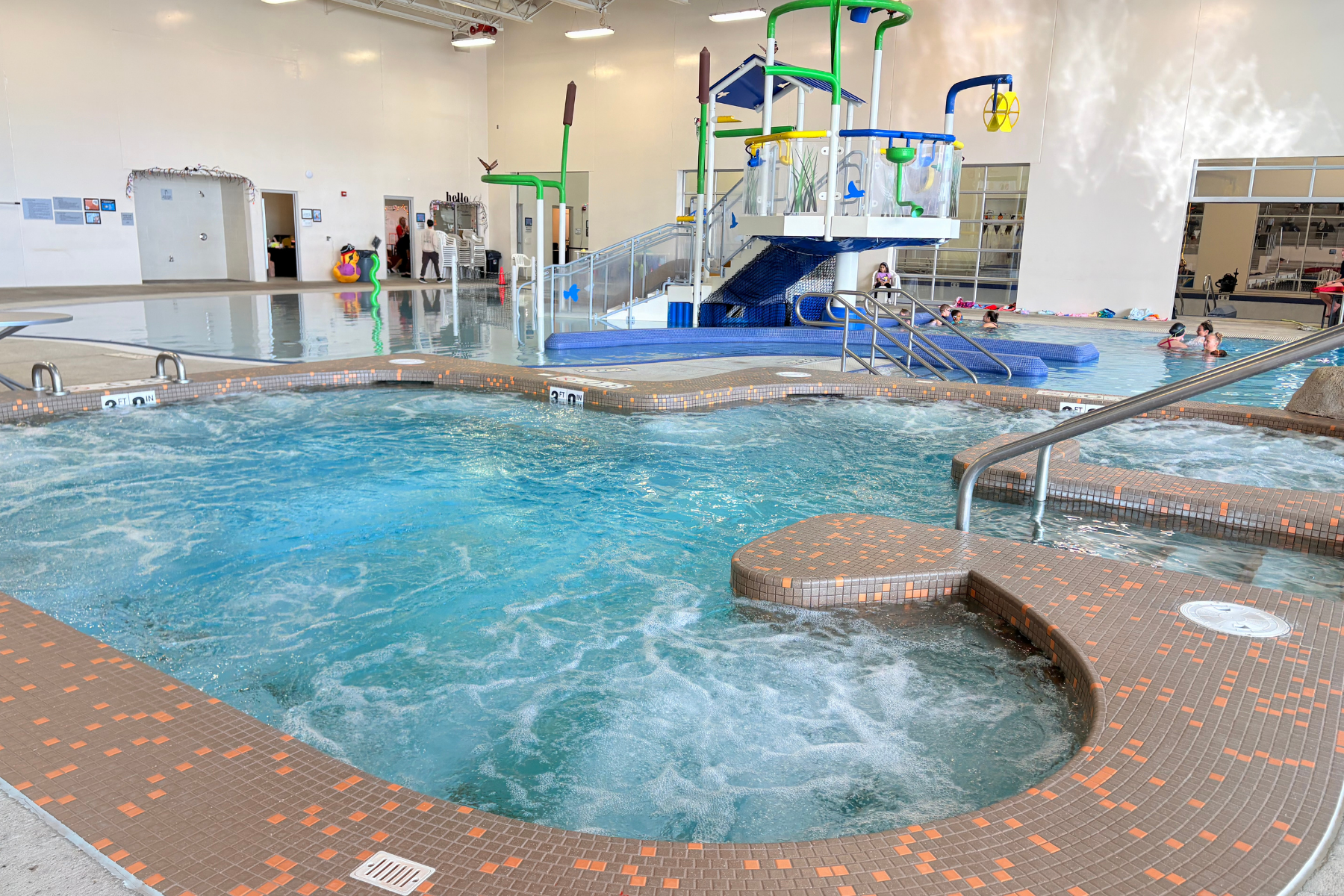 An indoor pool area featuring a hot tub, water slide, and kids swimming, with colorful play structures and bright lighting.