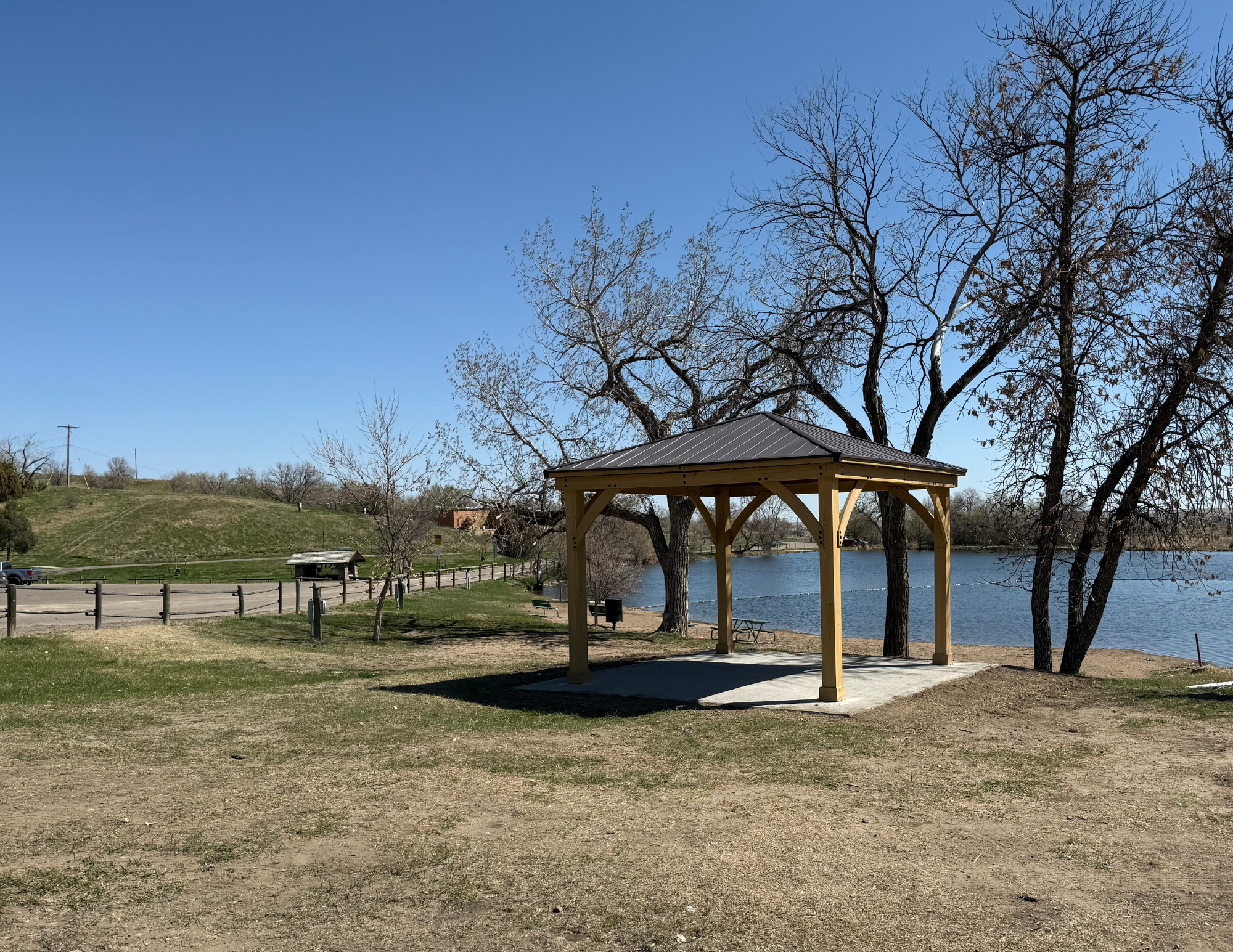 A gazebo by a lake with bare trees and a clear blue sky, and a fence leading to a distant building and hills.