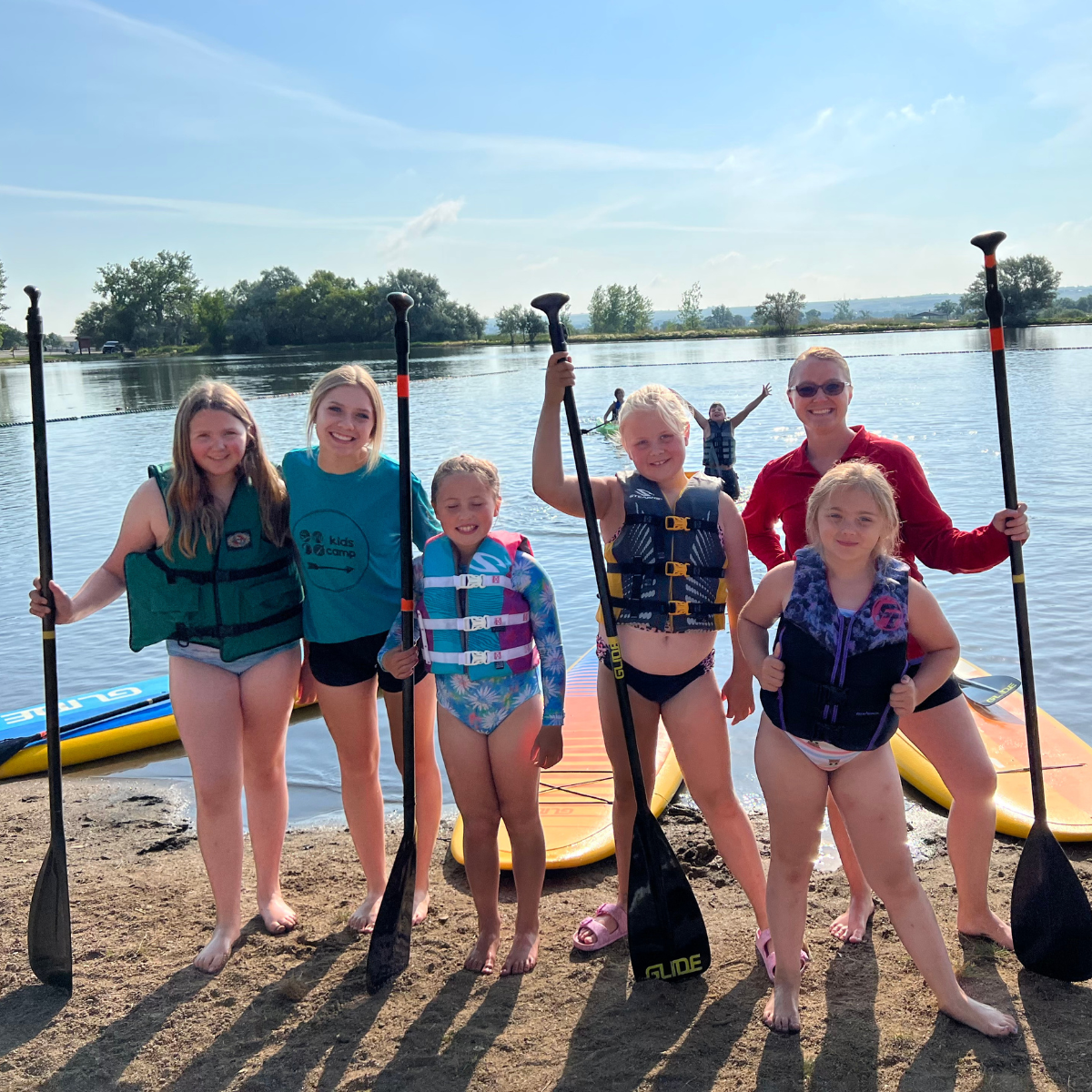 A group of children and an adult pose with paddles by a lake, all wearing life vests, enjoying a sunny day.