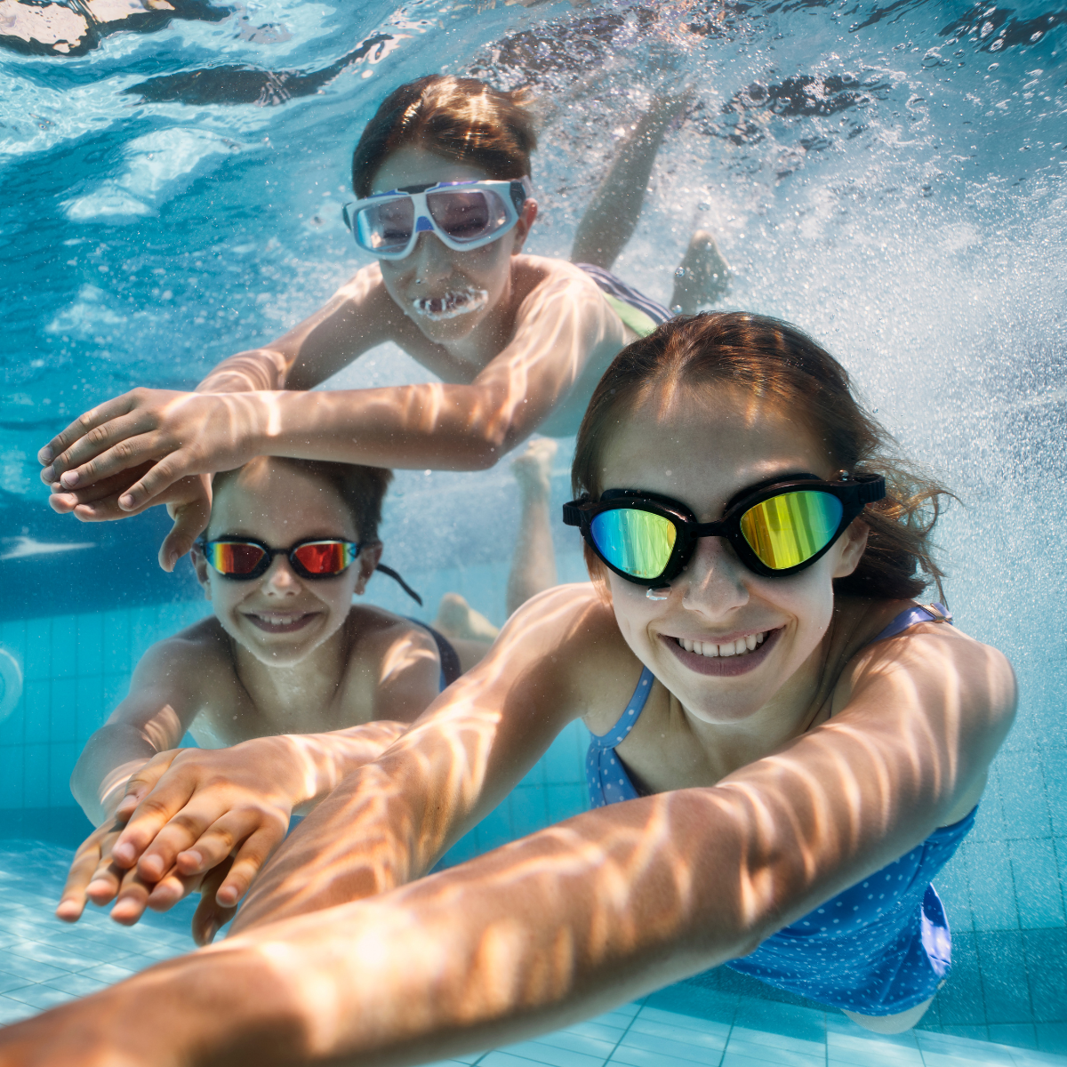 Three kids are playfully swimming underwater in a pool, wearing goggles and smiling amid dappled light and bubbles.