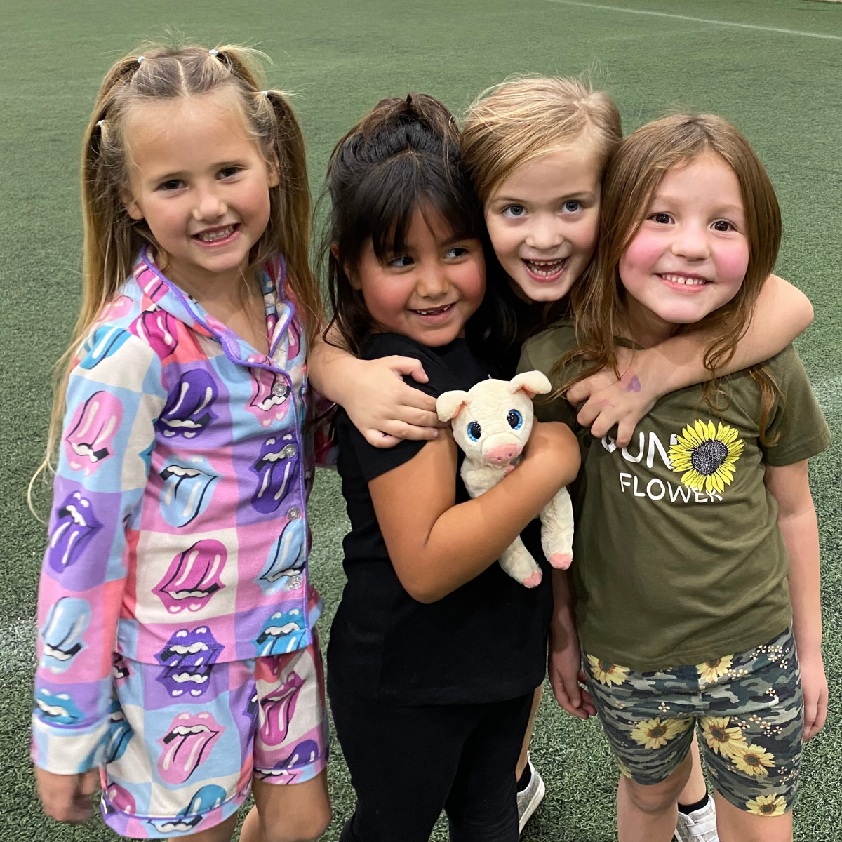A group of four smiling girls, wearing playful outfits, pose together, one holding a stuffed animal. A fun and cheerful moment!