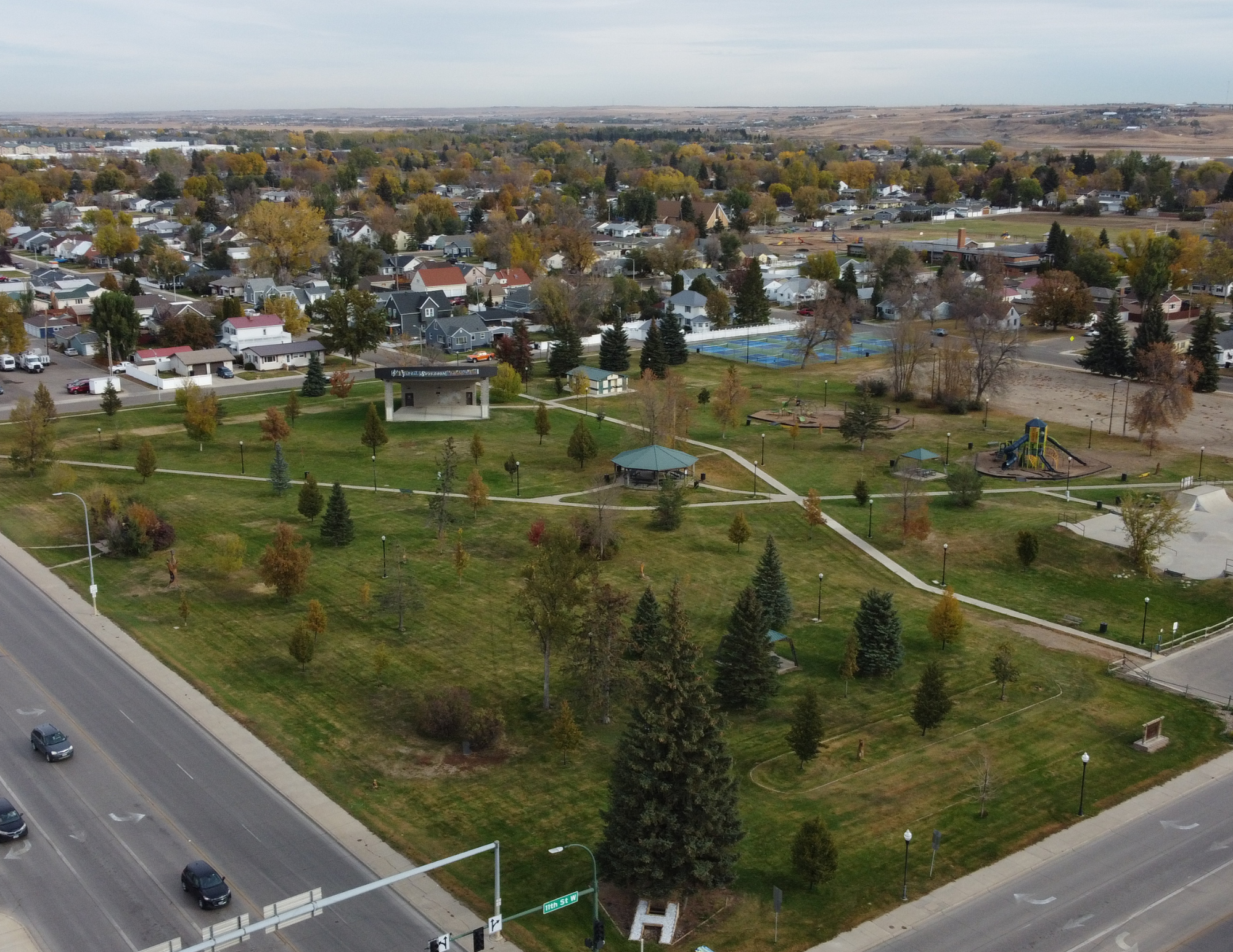 Aerial view of a town with a park, trees, pathways, a playground, and surrounding roads with cars.