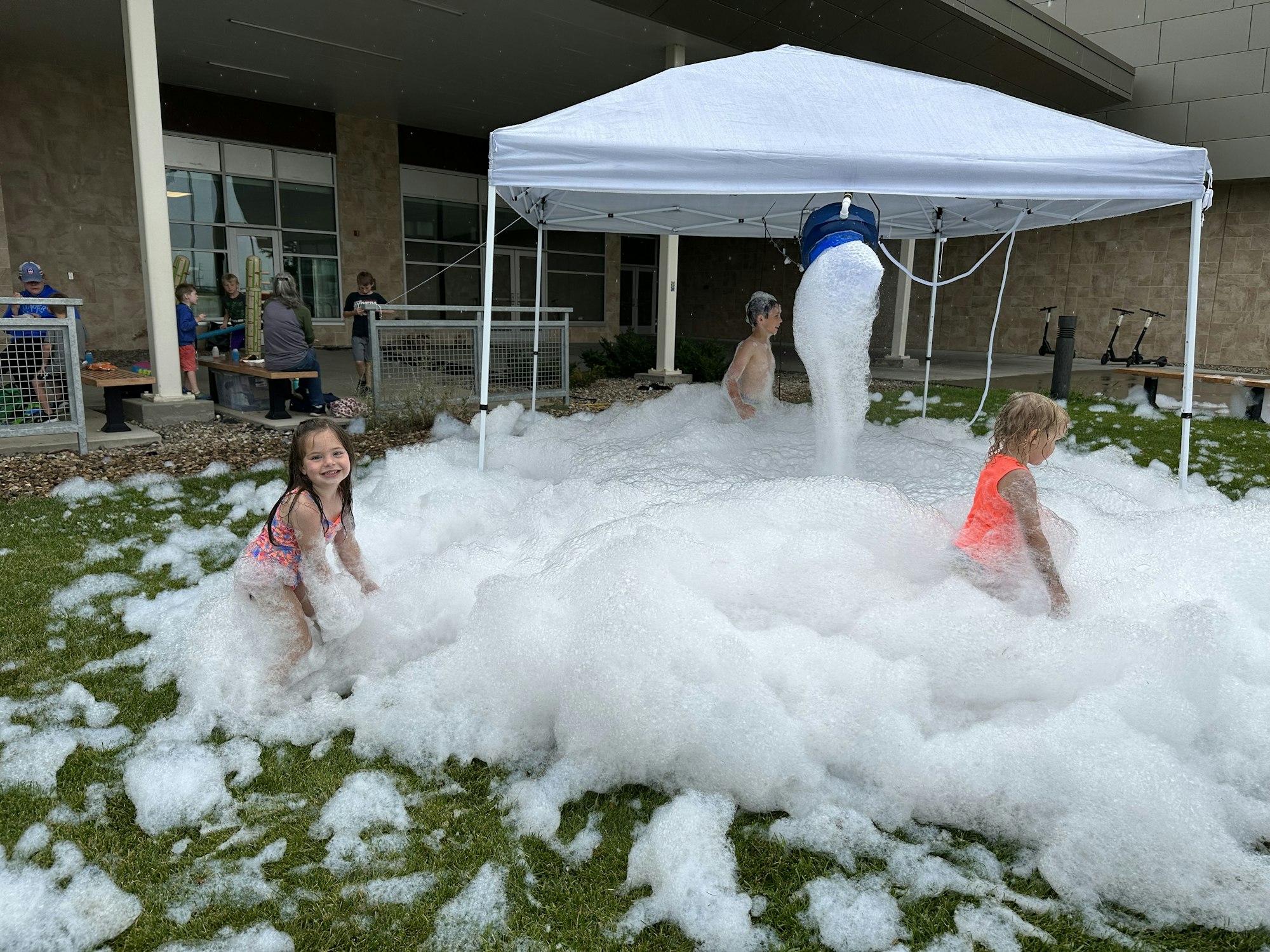 Children play in a foam pool under a canopy, with a foam machine overhead and bystanders nearby.