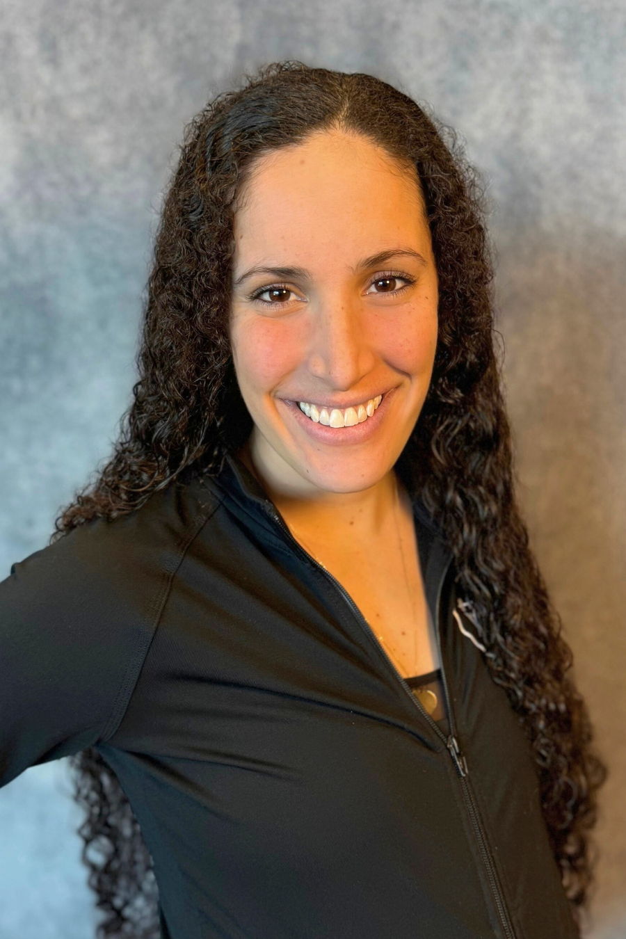 A person with long curly hair smiling, wearing a black zip-up top, with a blurred background.