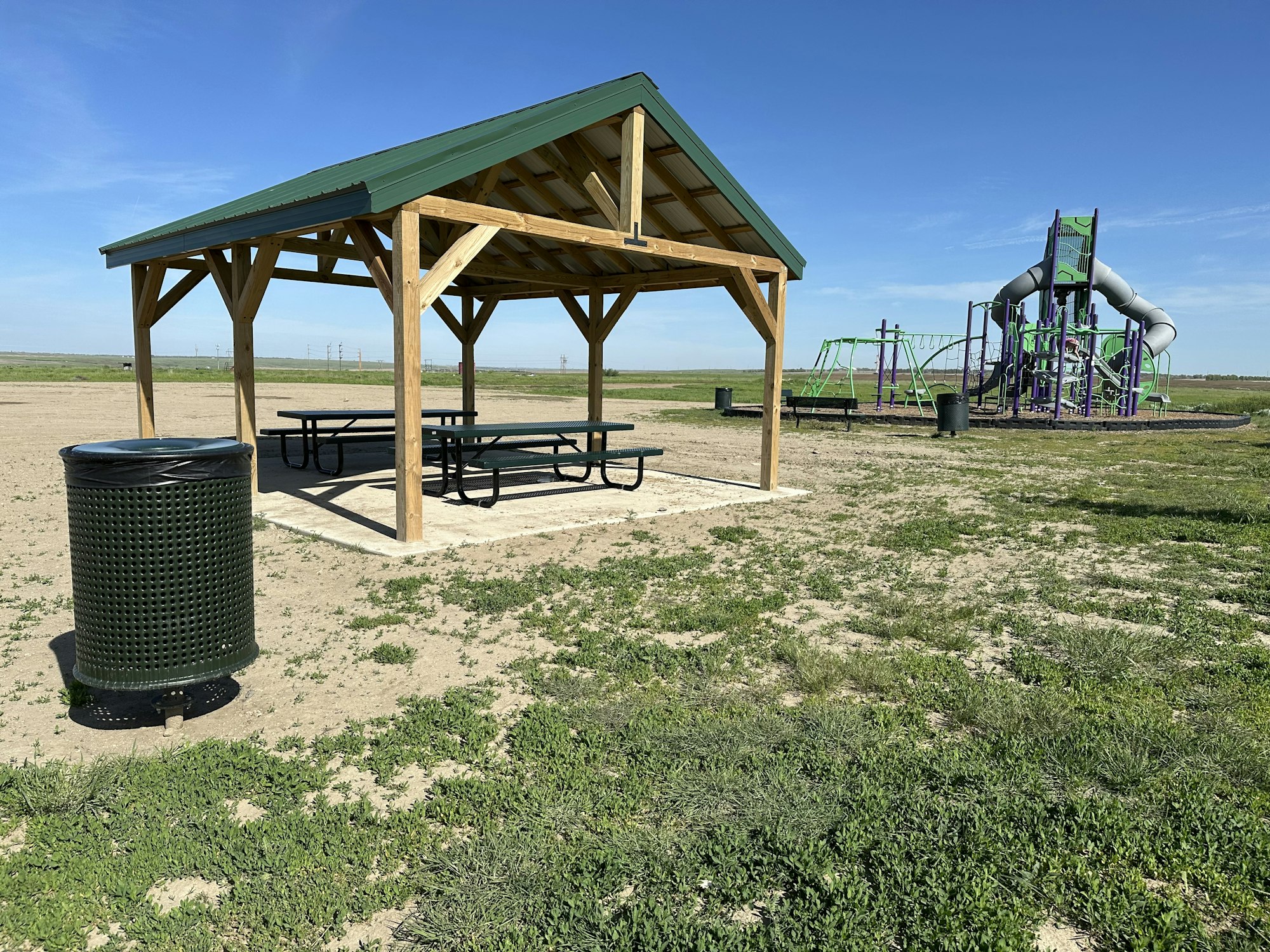 A park with a sheltered picnic area, trash bins, and a playground with slides in a grassy field under a clear sky.