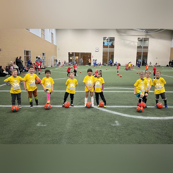 Children in yellow "Soccer" shirts stand on an indoor field with orange soccer balls.