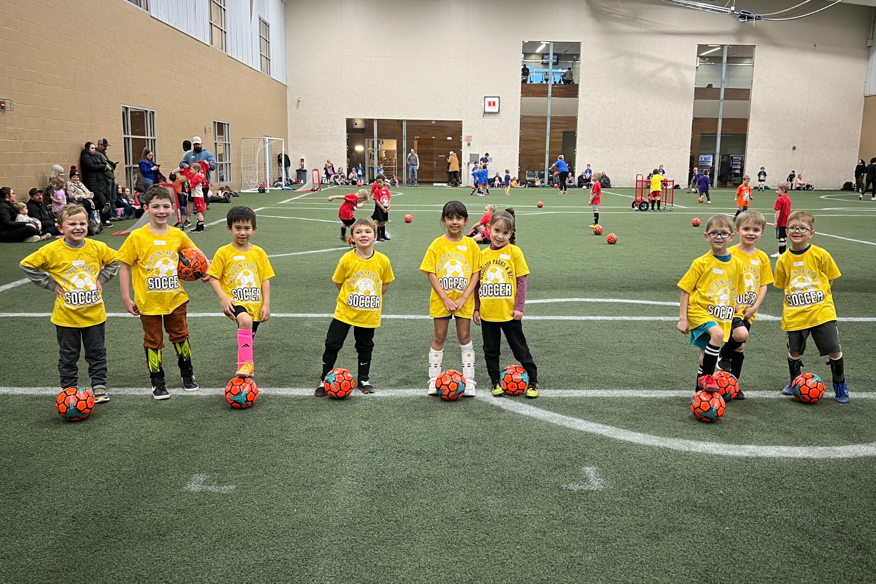 Children in yellow "Soccer" shirts stand on an indoor field with orange soccer balls.