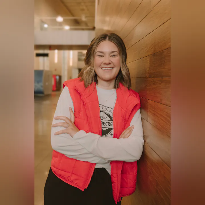 A smiling person in a red vest stands against a wooden wall, arms crossed, in a warm indoor setting.