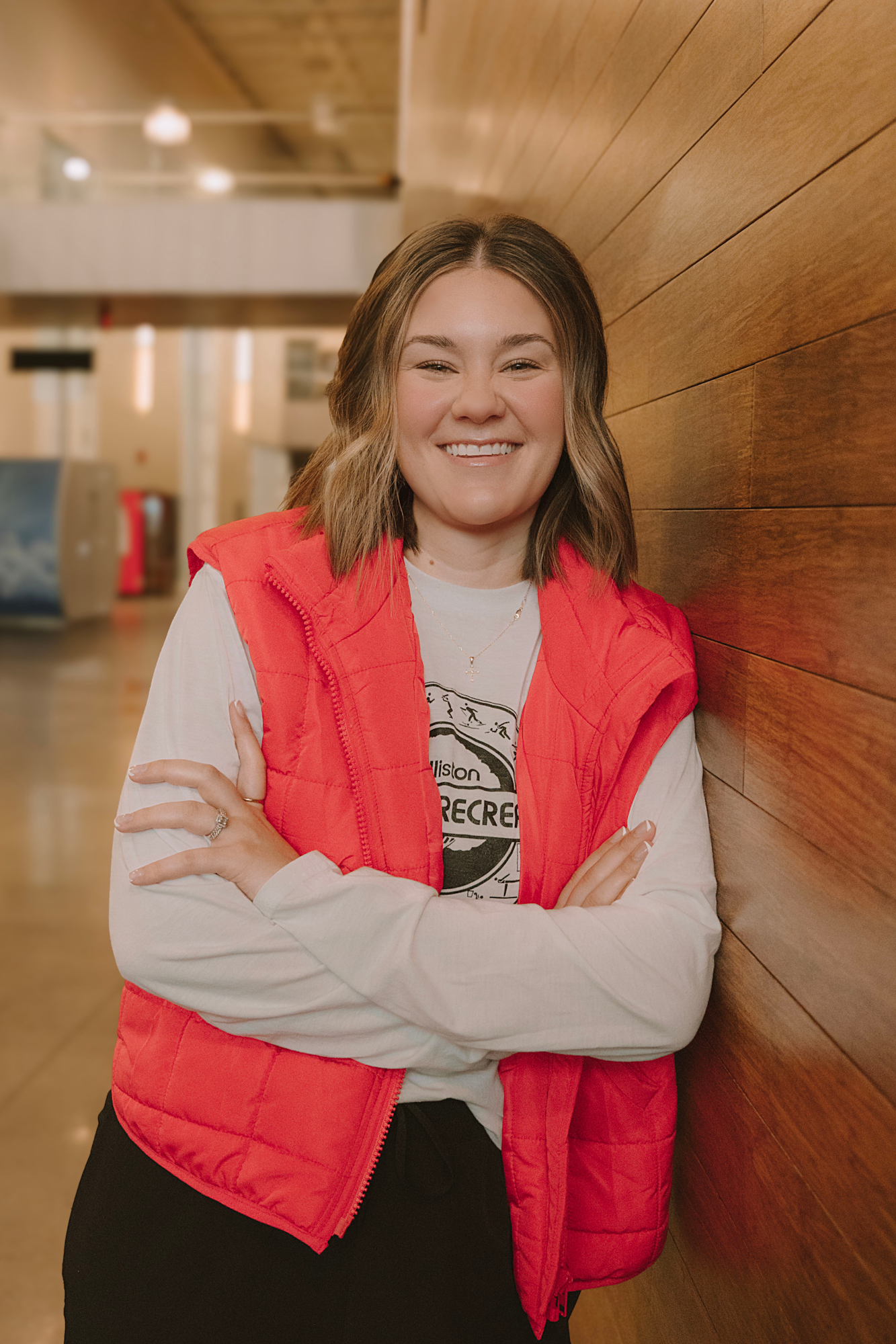 A smiling person in a red vest stands against a wooden wall, arms crossed, in a warm indoor setting.