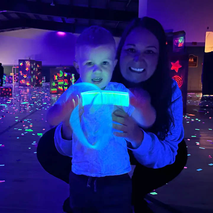 A child holding a glowing toy smiles with an adult in a colorful, festive environment, illuminated by black lights.