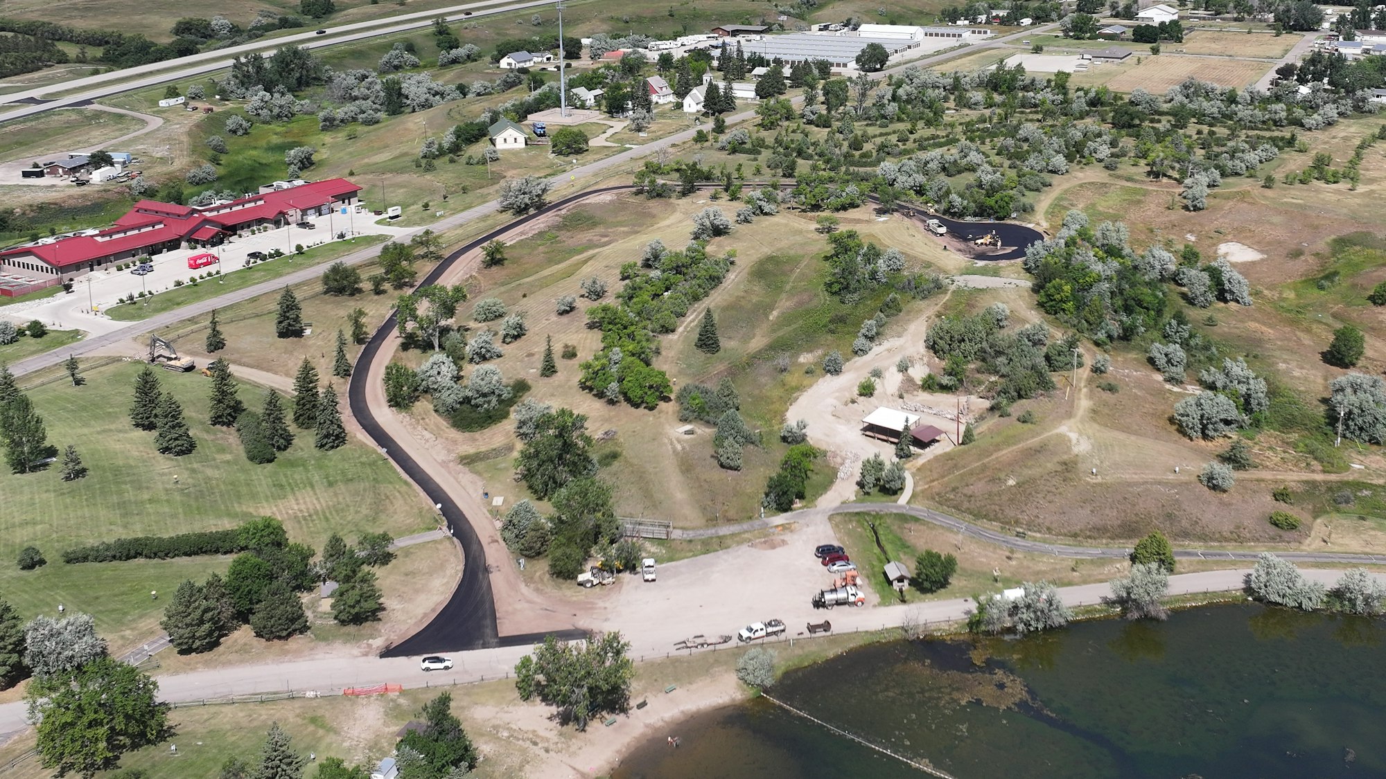 An aerial view of a green landscape with buildings, roads, and a body of water, featuring trees and a construction area.