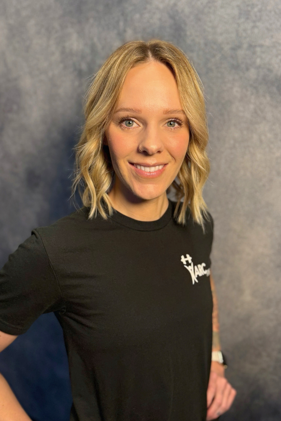 A person with blonde hair and a nose ring, smiling, wearing a black shirt with a white logo, standing against a gray background.