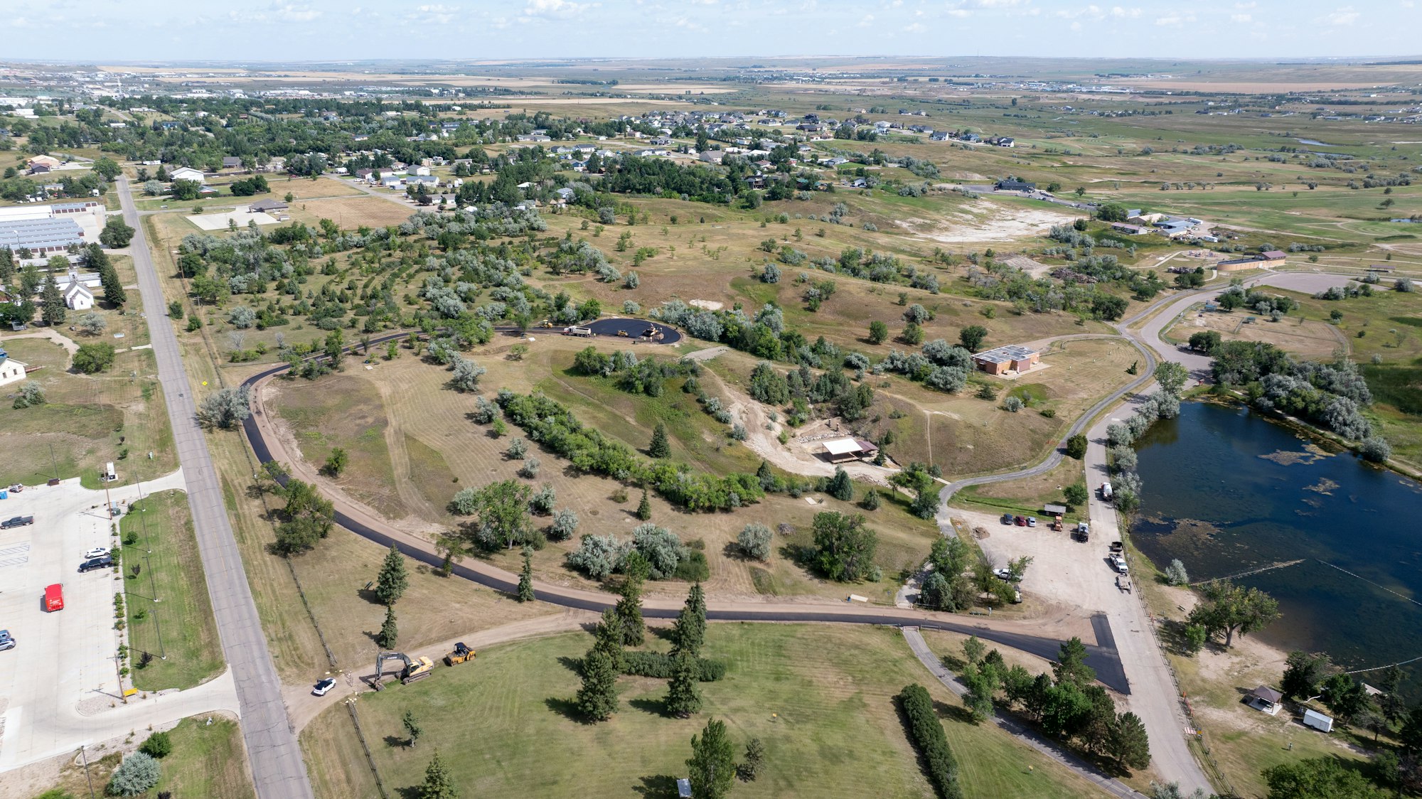 Aerial view of a rural area with roads, scattered buildings, green spaces, and a body of water, surrounded by open fields.
