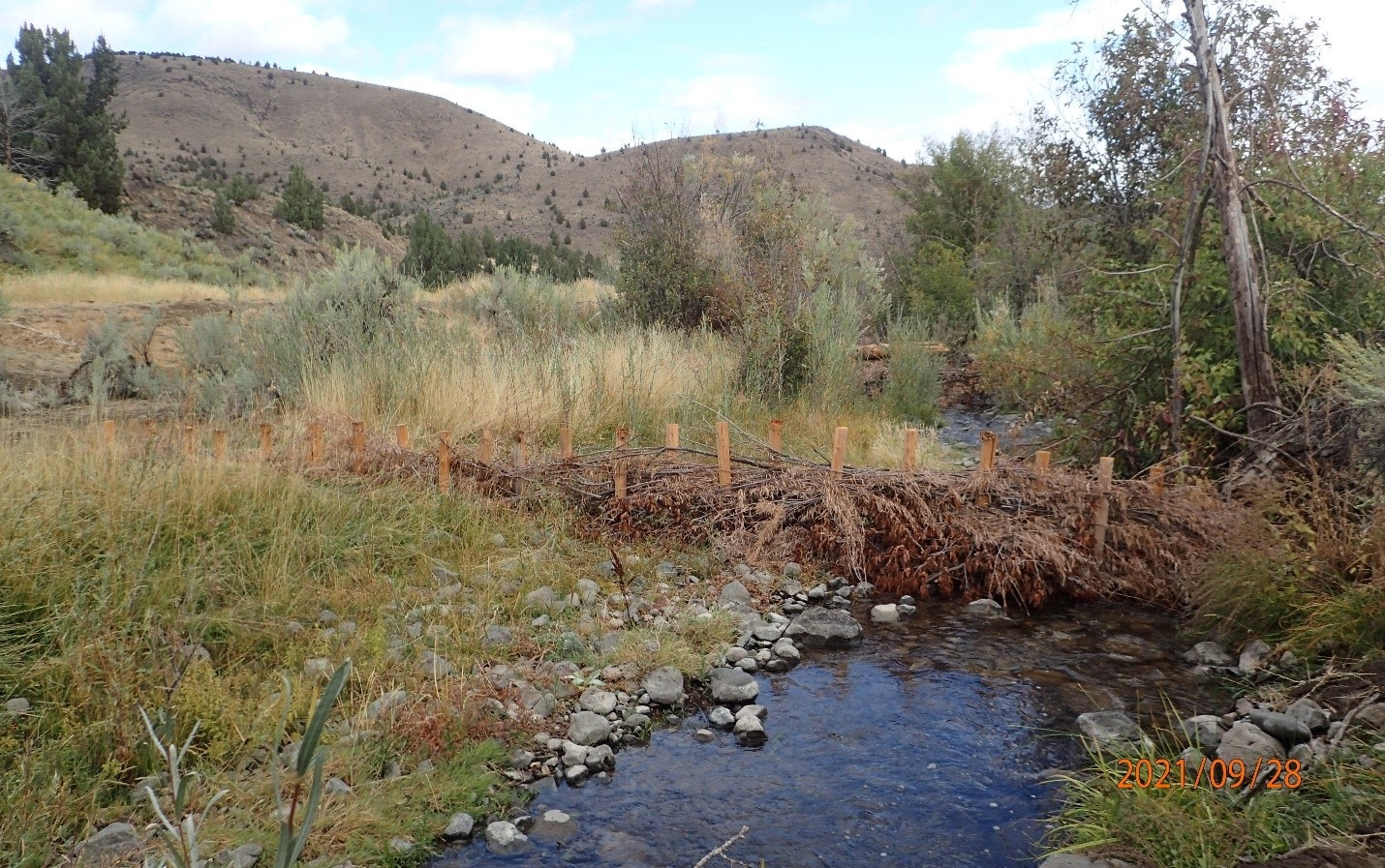 A stream with a beaver dam in a grassy landscape with hills in the background and a date stamp "2021/09/28."