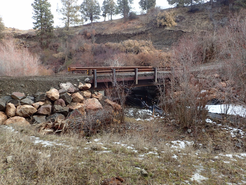 Rustic bridge over stream, rocky embankment, sparse vegetation, and light snow.
