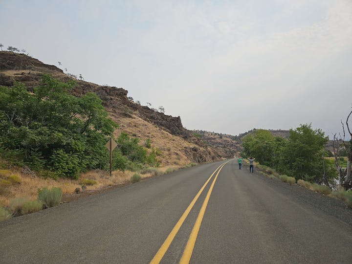 A road with double yellow lines leading through a rocky landscape with two people walking alongside and shrubs around.