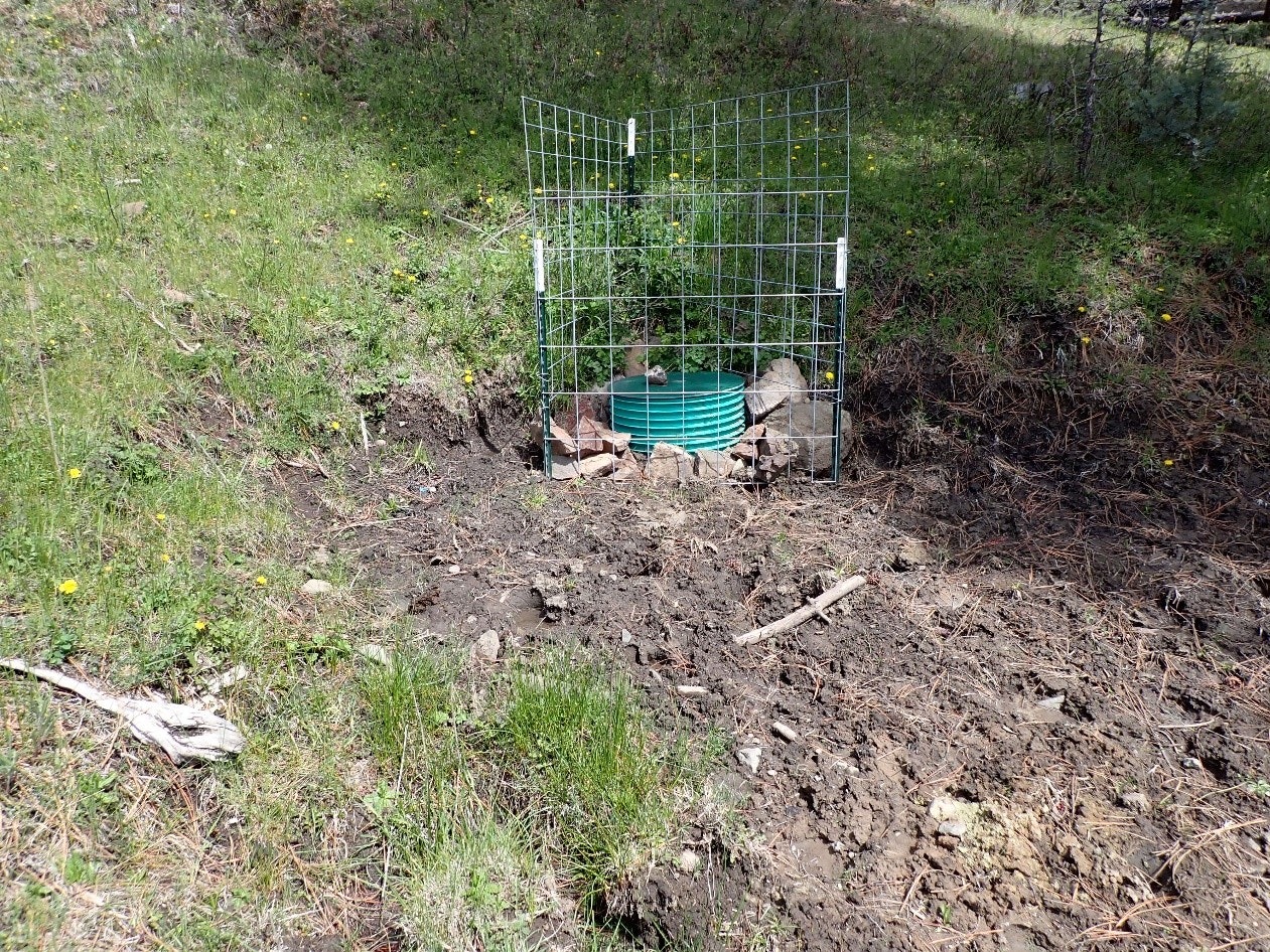 A green barrel within a wire fence, surrounded by disturbed soil and vegetation.