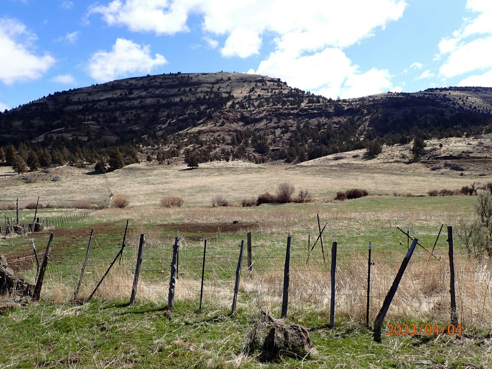 Rustic fence line with grassy field and hillside against a blue sky with clouds.