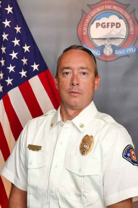 The image features a uniformed fire department official posing against a backdrop with an American flag and department logo.