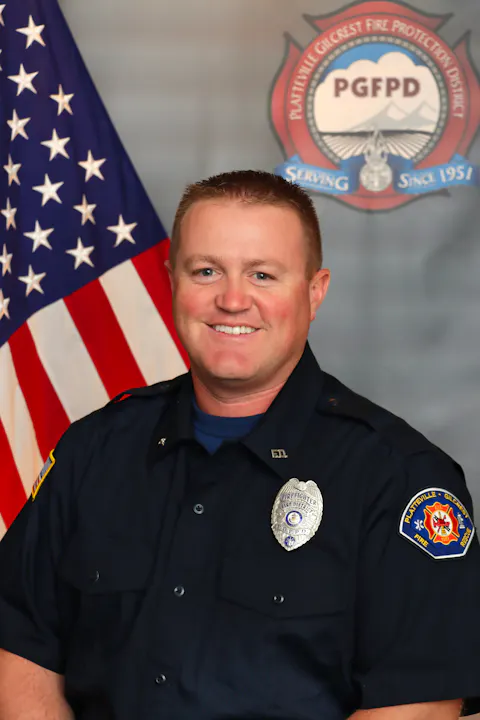 A smiling firefighter in uniform poses in front of an American flag and a fire department badge.