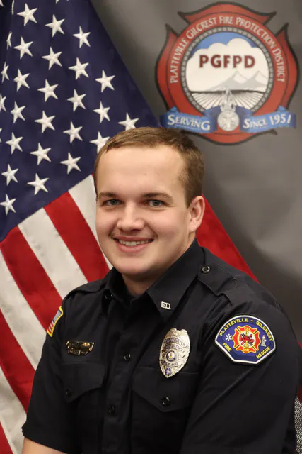 A smiling individual in a fire department uniform poses in front of a flag and an organizational emblem.