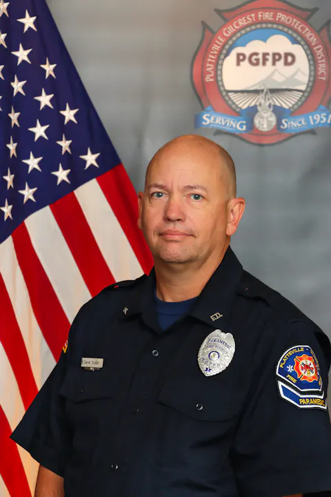 The image shows a paramedic in uniform, standing in front of an American flag and a fire protection district emblem.