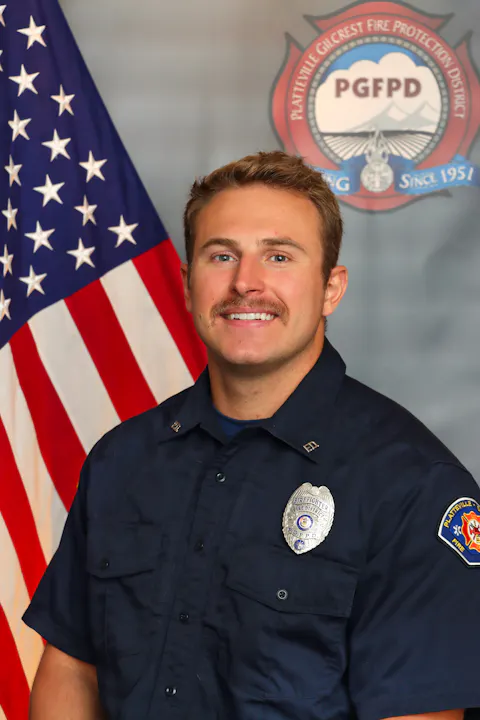 A firefighter in uniform smiles for a portrait, with an American flag and the logo of the fire protection district in the background.