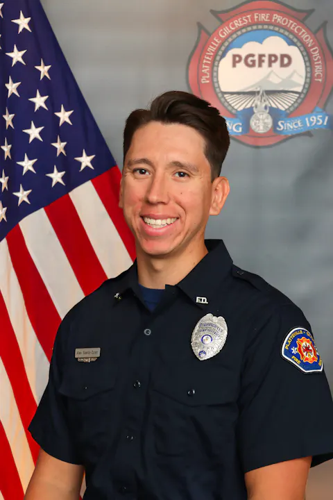 A firefighter in uniform smiles in front of an American flag and a fire department emblem.