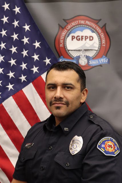 A man in a fire department uniform poses for a photo with an American flag and a department emblem in the background.