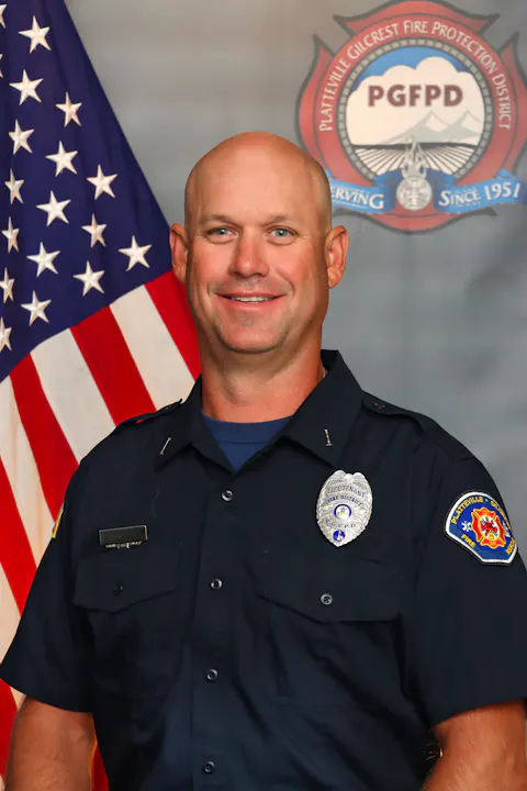 The image shows a smiling male officer in a uniform, standing in front of an American flag and a fire protection district emblem.