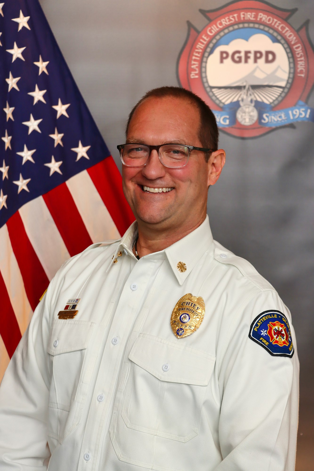 A smiling chief firefighter in a white uniform, with an American flag and a fire department emblem in the background.