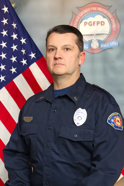A uniformed firefighter posing in front of an American flag and a fire department emblem.