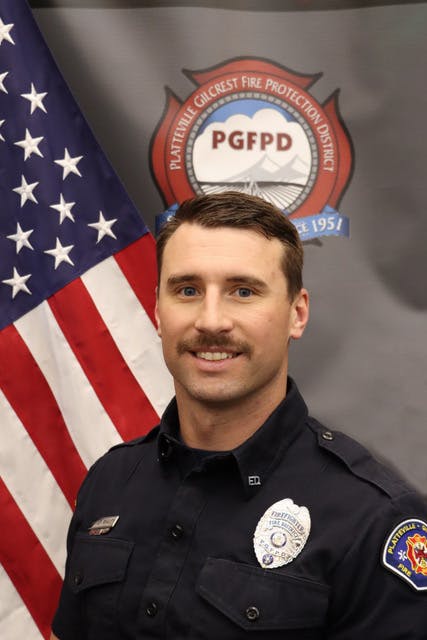 A firefighter in uniform poses for a portrait next to an American flag and a fire protection district emblem.