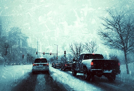 Vehicles stopped at a traffic light on a snowy road with trees and snowflakes visible.