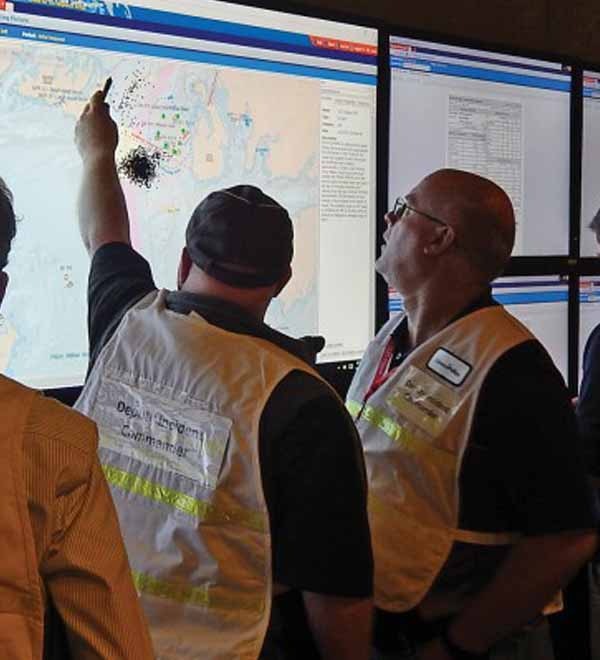 People in vests examine data on large monitors, one pointing at the screen.