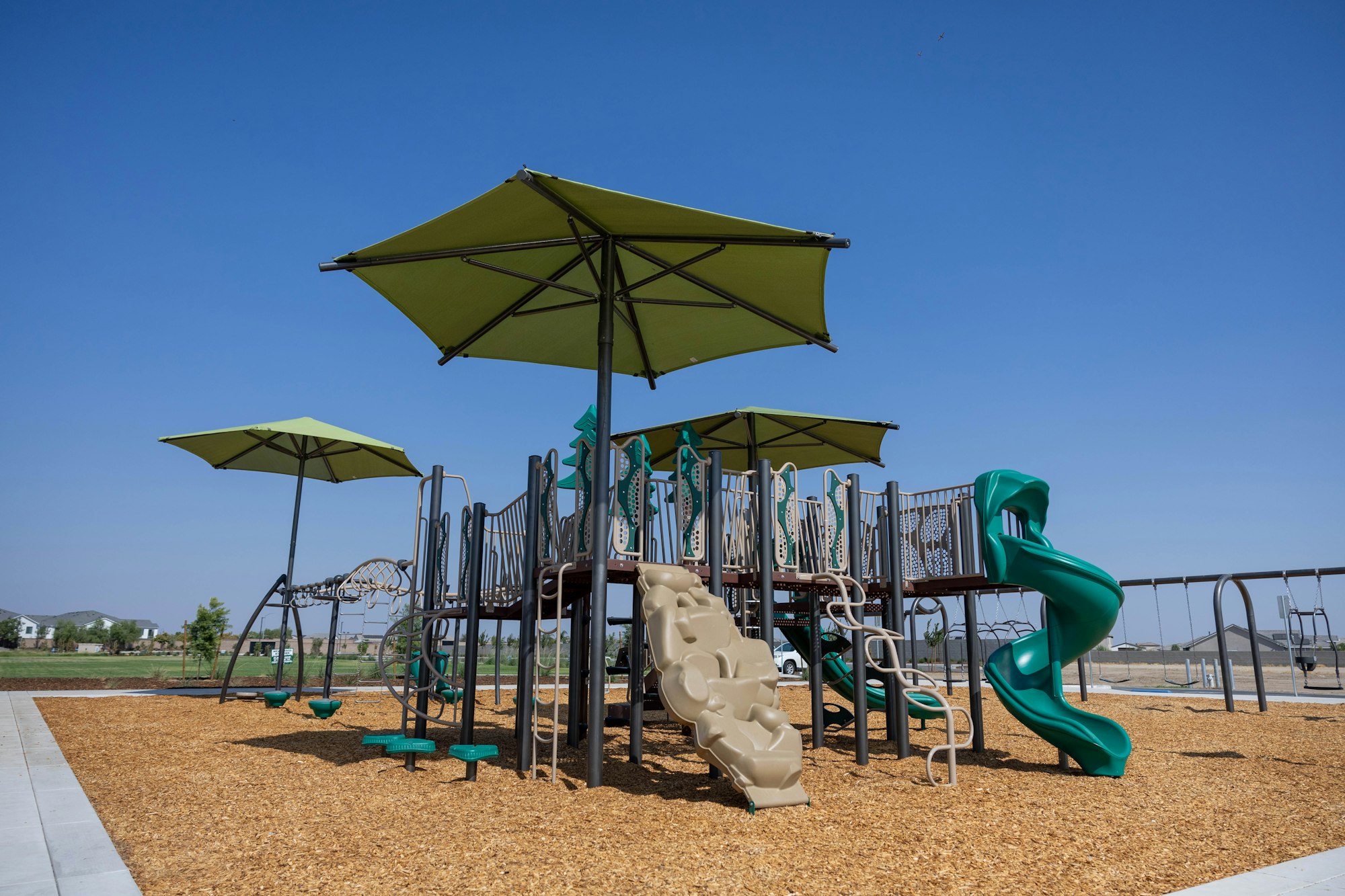 Outdoor playground with slides, swings, and climbers under green shade structures, set on woodchip ground.