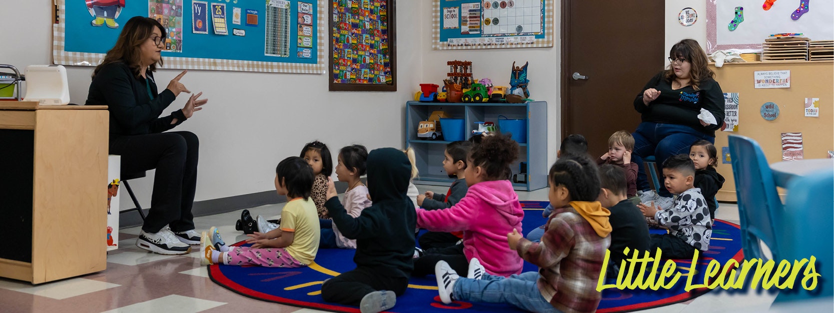 A teacher uses sign language with kids on a rug while another adult observes in a classroom titled "Little Learners."