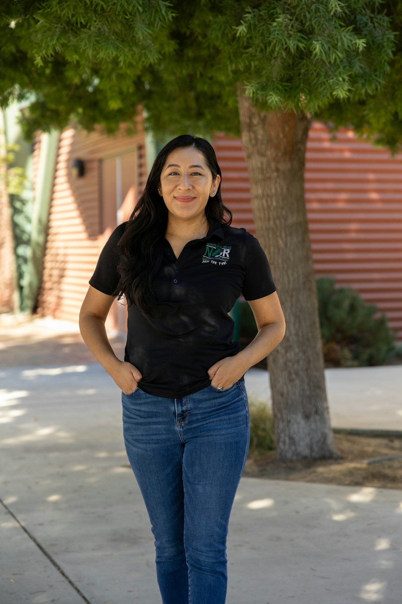 A person smiling, standing outdoors under a tree, wearing a black polo shirt and blue jeans.