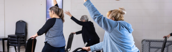 People in a fitness class holding chairs for balance, performing stretching exercises indoors.