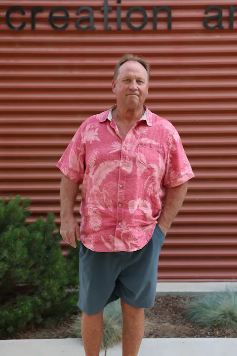 A man stands in front of a corrugated wall, wearing a pink floral shirt and gray shorts, with plants nearby.