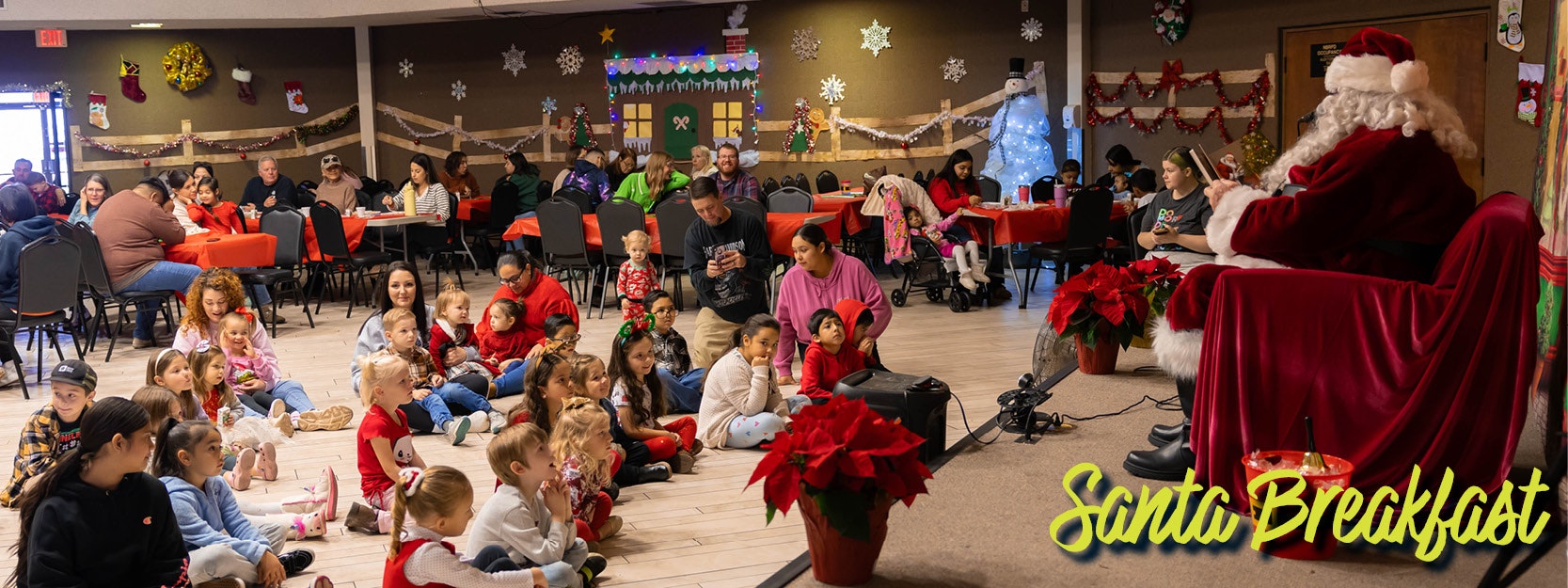 Children gathered for a Santa Breakfast in a festive room with decorations and poinsettias, while Santa sits on stage.