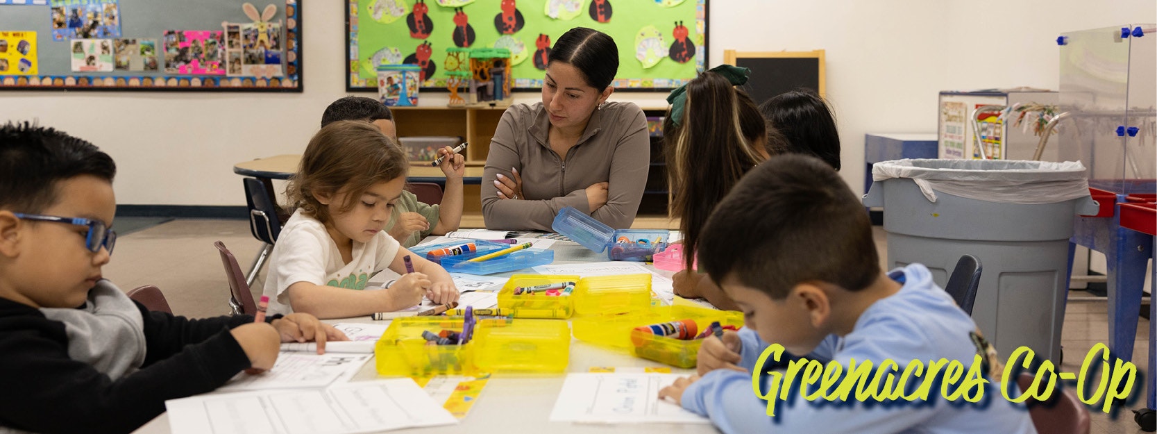 A teacher and young students coloring at a table in a classroom labeled "Greenacres Co-Op."