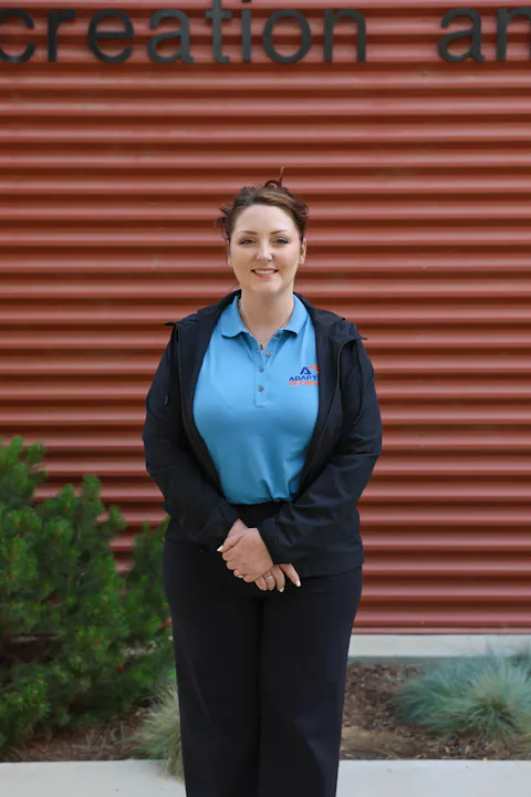 A woman stands smiling in front of a corrugated, red background with greenery nearby, wearing a blue polo and black jacket.