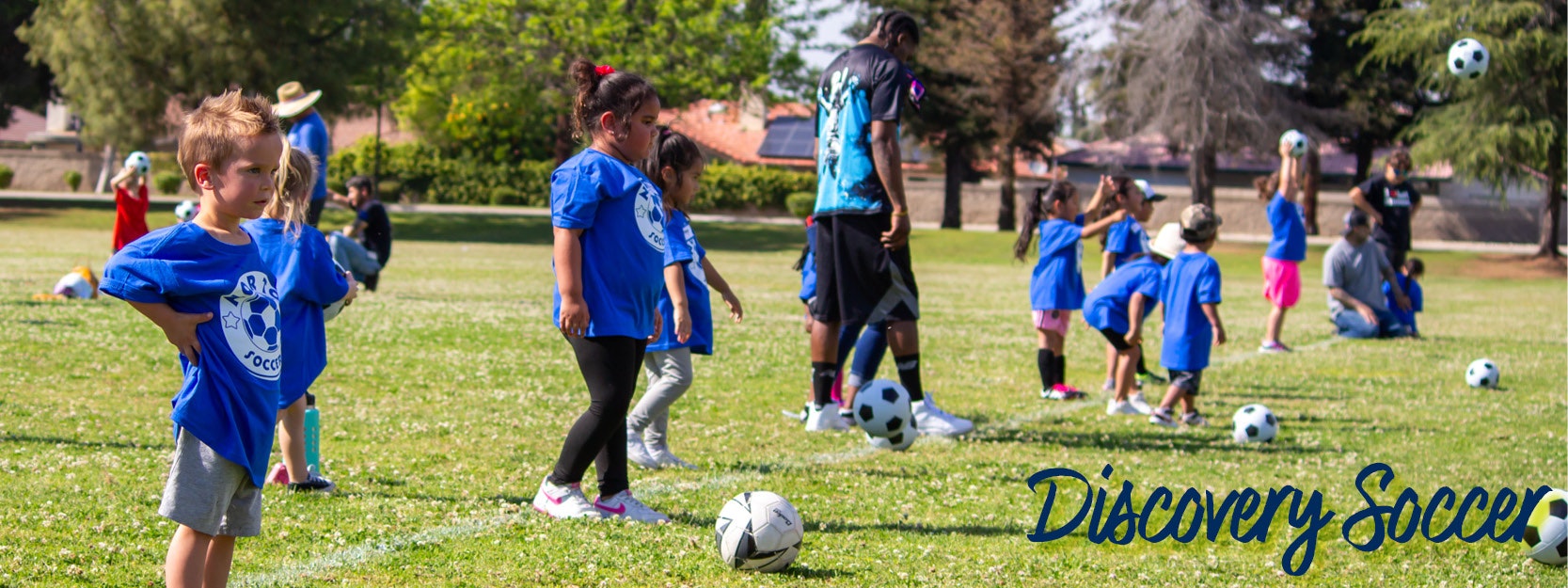 Children in blue shirts playing soccer on a field with adults supervising. Text reads "Discovery Soccer."