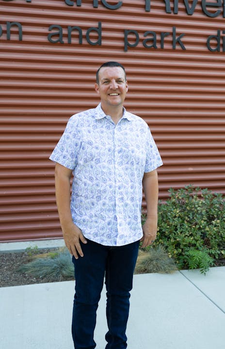 A smiling man in a patterned shirt and jeans standing in front of a building with signage.