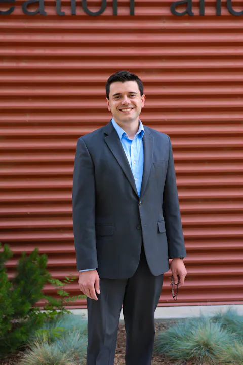 A man in a suit smiles while standing against a textured red wall, with greenery around him.